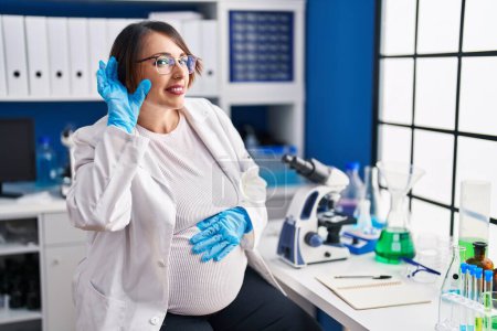 Téléchargez les photos : Pregnant woman working at scientist laboratory smiling with hand over ear listening an hearing to rumor or gossip. deafness concept. - en image libre de droit