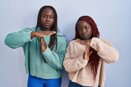 Foto de Dos mujeres africanas de pie sobre fondo azul haciendo un gesto de tiempo fuera con las manos, rostro frustrado y serio - Imagen libre de derechos