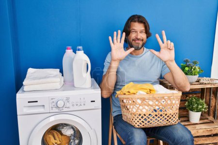 Téléchargez les photos : Bel homme d'âge moyen attendant la lessive montrant et pointant vers le haut avec les doigts numéro huit tout en souriant confiant et heureux. - en image libre de droit