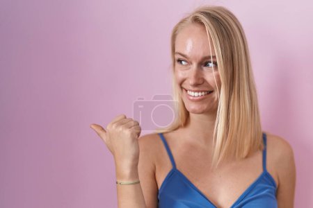 Téléchargez les photos : Jeune femme caucasienne debout sur fond rose souriant avec le visage heureux regardant et pointant vers le côté avec le pouce vers le haut. - en image libre de droit