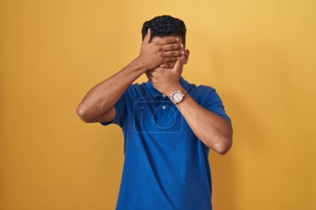 Photo for Young hispanic man standing over yellow background covering eyes and mouth with hands, surprised and shocked. hiding emotion - Royalty Free Image