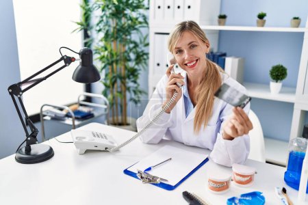 Téléchargez les photos : Young blonde woman wearing dentist uniform talking on the telephone holding xray at clinic - en image libre de droit