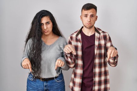Foto de Young hispanic couple standing over white background pointing down looking sad and upset, indicating direction with fingers, unhappy and depressed. - Imagen libre de derechos