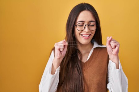 Photo for Young brunette woman standing over yellow background wearing glasses excited for success with arms raised and eyes closed celebrating victory smiling. winner concept. - Royalty Free Image
