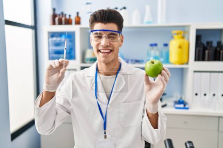 Photo for Young hispanic man working at scientist laboratory holding apple winking looking at the camera with sexy expression, cheerful and happy face. - Royalty Free Image