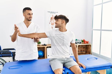 Téléchargez les photos : Deux hommes hispaniques physiothérapeute et patient ayant session de réadaptation étirant le bras à la clinique - en image libre de droit