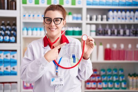 Photo for Young hispanic woman with red hair working at pharmacy drugstore using stethoscope smiling happy pointing with hand and finger - Royalty Free Image