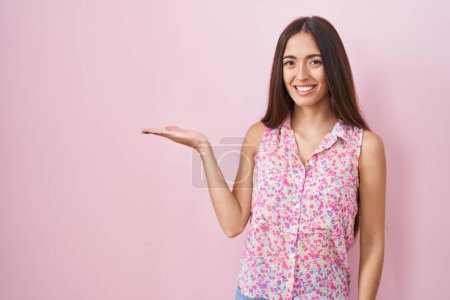 Foto de Mujer hispana joven con el pelo largo de pie sobre fondo rosa sonriente alegre presentando y señalando con la palma de la mano mirando a la cámara. - Imagen libre de derechos