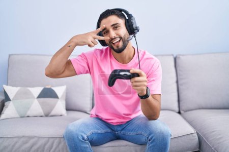 Téléchargez les photos : Jeune homme hispanique jouant contrôleur de tenue de jeu vidéo assis sur le canapé faisant symbole de paix avec les doigts sur le visage, souriant joyeux montrant la victoire - en image libre de droit