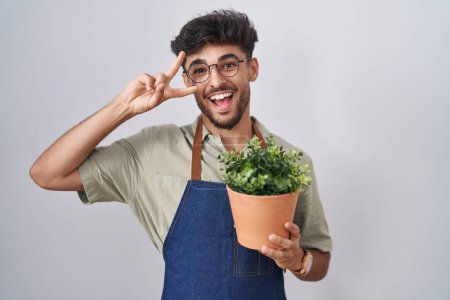 Téléchargez les photos : Homme arabe avec barbe tenant pot de plante verte faisant symbole de paix avec les doigts sur le visage, souriant joyeux montrant la victoire - en image libre de droit
