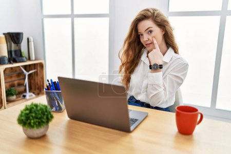 Photo for Young caucasian woman working at the office using computer laptop pointing to the eye watching you gesture, suspicious expression - Royalty Free Image