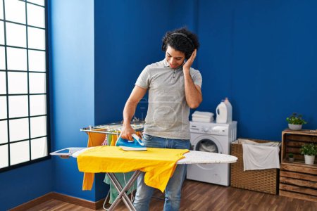 Foto de Joven hispano escuchando música planchando ropa en la lavandería - Imagen libre de derechos