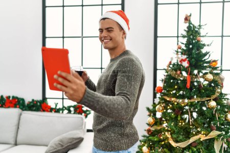 Téléchargez les photos : Young hispanic man make selfie by the touchpad drinking wine standing by christmas tree at home - en image libre de droit