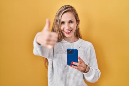 Photo for Young blonde woman using smartphone typing message approving doing positive gesture with hand, thumbs up smiling and happy for success. winner gesture. - Royalty Free Image