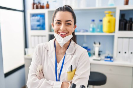 Photo for Young beautiful hispanic woman scientist wearing medical mask sitting with arms crossed gesture at laboratory - Royalty Free Image