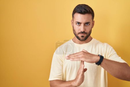 Photo for Handsome hispanic man standing over yellow background doing time out gesture with hands, frustrated and serious face - Royalty Free Image