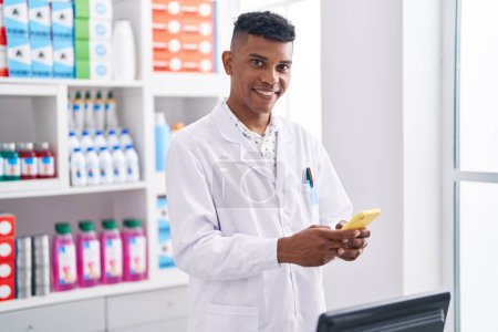 Téléchargez les photos : Jeune pharmacien homme latin souriant confiant en utilisant smartphone à la pharmacie - en image libre de droit
