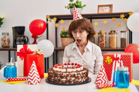Foto de Middle age woman celebrating birthday holding big chocolate cake scared and amazed with open mouth for surprise, disbelief face - Imagen libre de derechos