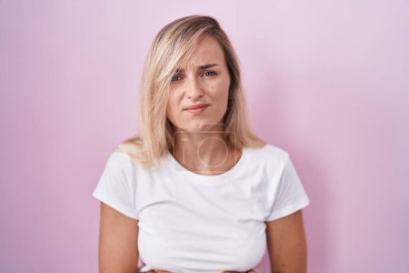 Foto de Young blonde woman standing over pink background with hand on stomach because indigestion, painful illness feeling unwell. ache concept. - Imagen libre de derechos