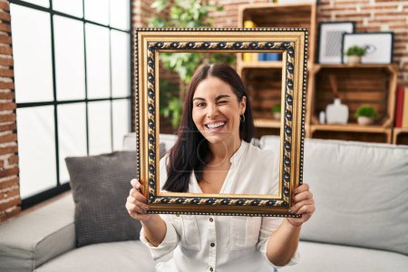 Photo for Young hispanic woman holding empty frame winking looking at the camera with sexy expression, cheerful and happy face. - Royalty Free Image