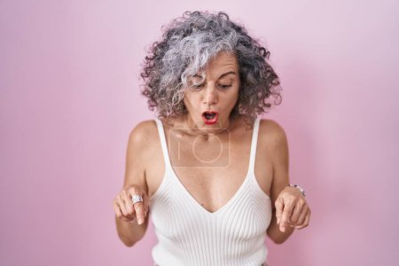 Photo for Middle age woman with grey hair standing over pink background pointing down with fingers showing advertisement, surprised face and open mouth - Royalty Free Image
