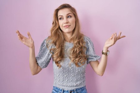 Téléchargez les photos : Beautiful blonde woman standing over pink background clueless and confused expression with arms and hands raised. doubt concept. - en image libre de droit