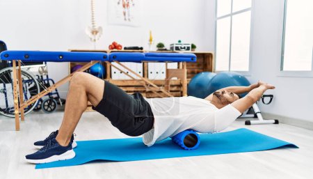 Photo for Young hispanic man patient having back rehab session using foam roller at clinic - Royalty Free Image