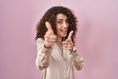Téléchargez les photos : Femme hispanique avec des cheveux bouclés debout sur fond rose pointant les doigts vers la caméra avec un visage heureux et drôle. bonne énergie et vibes. - en image libre de droit
