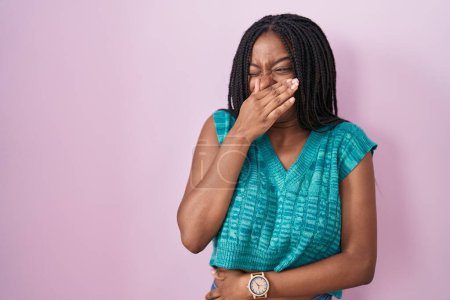 Téléchargez les photos : Young african american with braids standing over pink background smelling something stinky and disgusting, intolerable smell, holding breath with fingers on nose. bad smell - en image libre de droit
