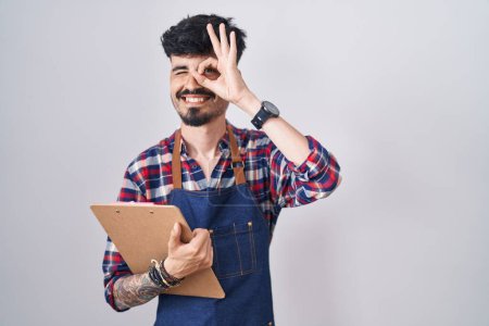 Foto de Young hispanic man with beard wearing waiter apron holding clipboard doing ok gesture with hand smiling, eye looking through fingers with happy face. - Imagen libre de derechos