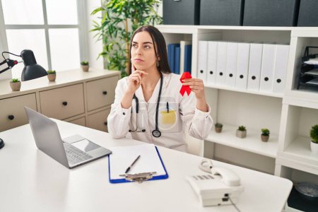 Photo for Hispanic doctor woman holding support red ribbon serious face thinking about question with hand on chin, thoughtful about confusing idea - Royalty Free Image