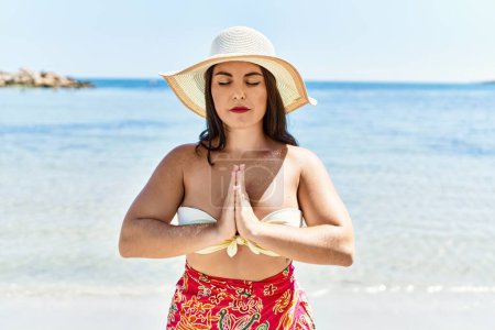 Téléchargez les photos : Jeune belle femme hispanique touriste portant un bikini et un chapeau d'été faisant de l'exercice de yoga au bord de la mer - en image libre de droit