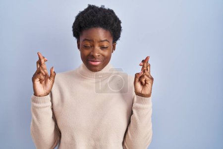 Téléchargez les photos : Femme afro-américaine debout sur fond bleu geste doigt croisé souriant avec espoir et les yeux fermés. chance et concept superstitieux. - en image libre de droit