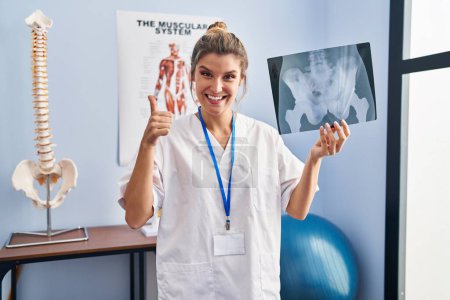 Photo for Young woman holding pelvis radiography smiling happy and positive, thumb up doing excellent and approval sign - Royalty Free Image