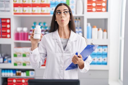 Photo for Young brunette woman working at pharmacy drugstore holding pills making fish face with mouth and squinting eyes, crazy and comical. - Royalty Free Image
