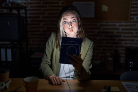 Foto de Blonde caucasian woman working at the office at night puffing cheeks with funny face. mouth inflated with air, crazy expression. - Imagen libre de derechos