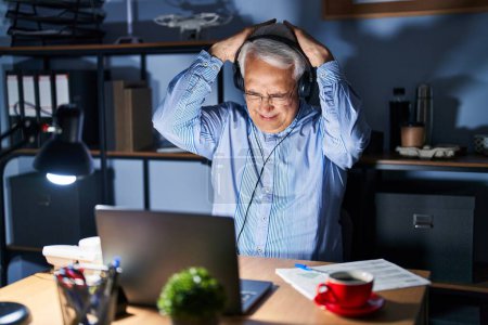 Foto de Hombre mayor hispano con auriculares de agente de centro de llamadas en la noche que sufre de dolor de cabeza desesperado y estresado debido al dolor y la migraña. manos en la cabeza. - Imagen libre de derechos
