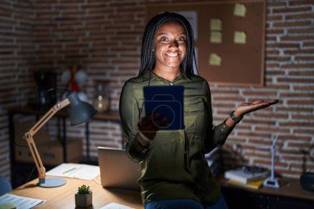 Téléchargez les photos : Young african american with braids working at the office at night smiling cheerful presenting and pointing with palm of hand looking at the camera. - en image libre de droit