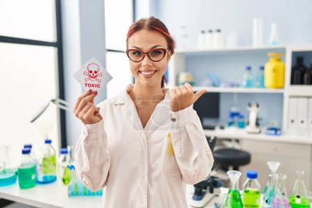 Téléchargez les photos : Jeune femme caucasienne travaillant au laboratoire scientifique tenant une étiquette toxique pointant le pouce vers le côté souriant heureux avec la bouche ouverte - en image libre de droit