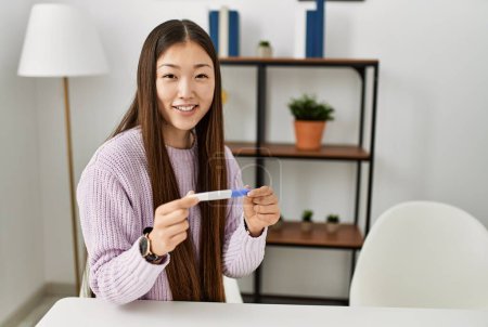 Foto de Joven chica china sosteniendo prueba de embarazo sentado en la mesa en casa. - Imagen libre de derechos
