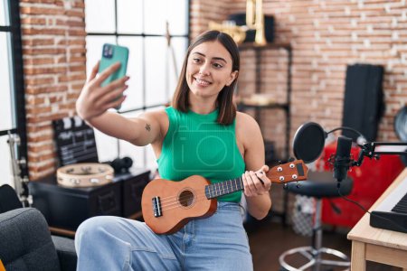 Photo for Young hispanic woman musician make selfie by the smartphone holding ukelele at music studio - Royalty Free Image