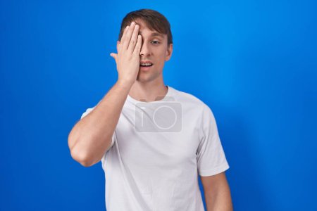 Photo for Caucasian blond man standing over blue background covering one eye with hand, confident smile on face and surprise emotion. - Royalty Free Image