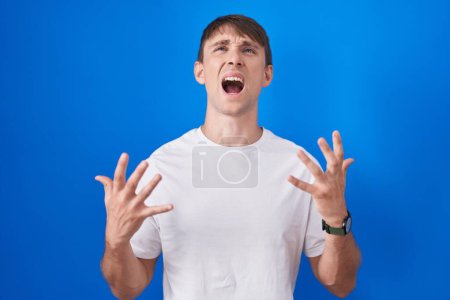 Foto de Caucasian blond man standing over blue background crazy and mad shouting and yelling with aggressive expression and arms raised. frustration concept. - Imagen libre de derechos