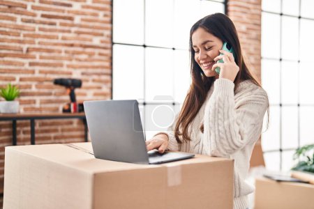 Photo for Young woman talking on the smartphone using laptop at new home - Royalty Free Image