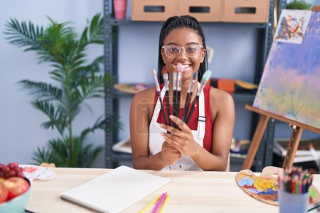 Foto de African american woman smiling confident holding paintbrushes at art studio - Imagen libre de derechos