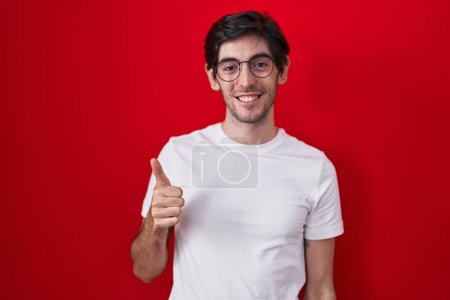 Téléchargez les photos : Jeune homme hispanique debout sur fond rouge faisant des pouces heureux geste avec la main. approuvant l'expression regardant la caméra montrant le succès. - en image libre de droit