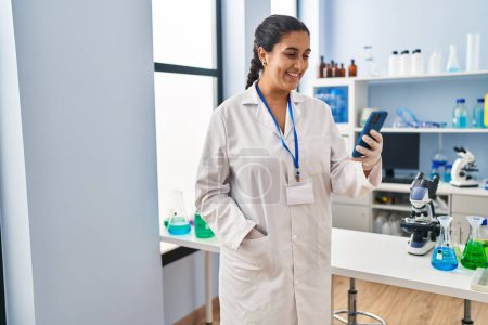 Téléchargez les photos : Jeune femme hispanique portant un uniforme de scientifique en utilisant un smartphone au laboratoire - en image libre de droit
