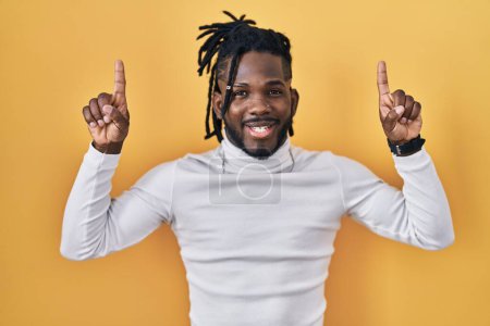 Foto de Hombre africano con rastas con jersey de cuello alto sobre fondo amarillo sonriendo asombrado y sorprendido y señalando con los dedos y los brazos levantados. - Imagen libre de derechos