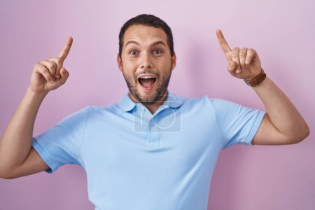 Foto de Hombre hispano de pie sobre fondo rosa sonriendo asombrado y sorprendido y señalando con los dedos y los brazos levantados. - Imagen libre de derechos