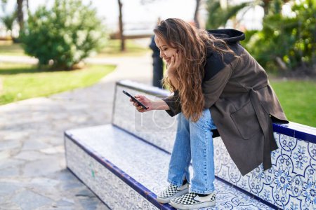 Téléchargez les photos : Young beautiful hispanic woman using smartphone sitting on bench at park - en image libre de droit